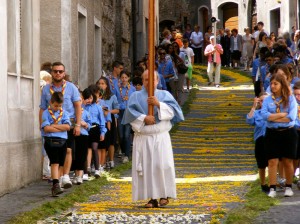 A priest is carrying a cross down the flowered streets of Cagli. Behind him are scouts. The scouts come early to help decorate the streets in honor of the procession.