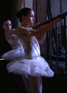 Two young ballerinas in white tutus watch patiently from backstage as the older dancers stretch and warm up for Sleeping Beauty, waiting for their moment to shine.