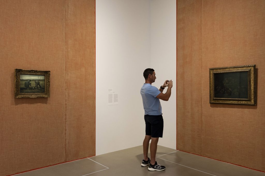 A man stands in a museum taking a photo of a painting on the wall.