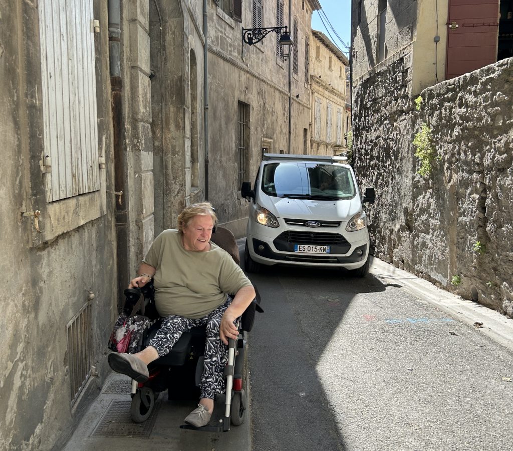 Woman in a wheelchair maneuvers through a narrow street in Arles as a car tries to pass her.