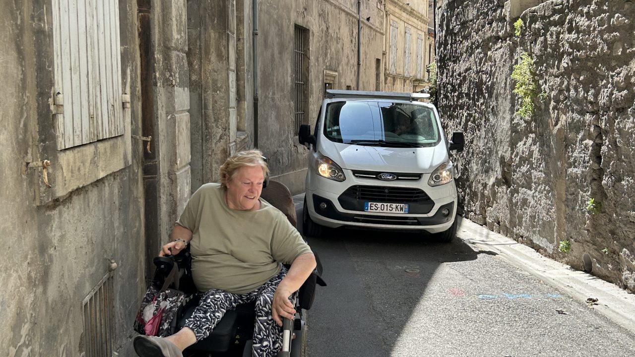 Woman in a wheelchair maneuvers through a narrow street in Arles as a car tries to pass her.
