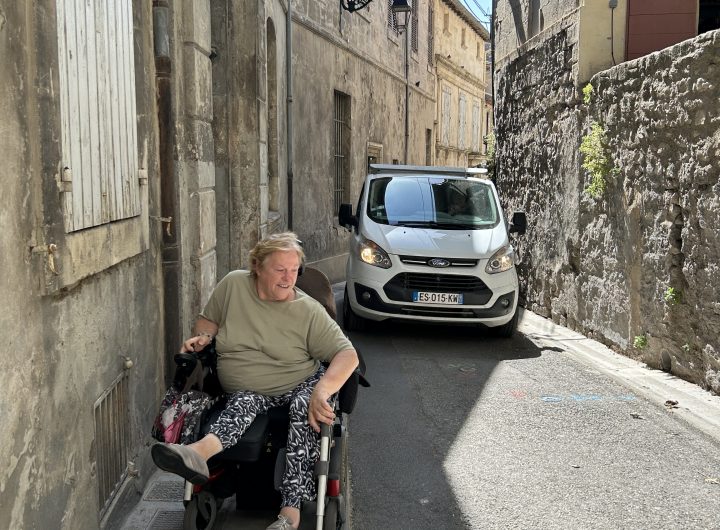 Woman in a wheelchair maneuvers through a narrow street in Arles as a car tries to pass her.