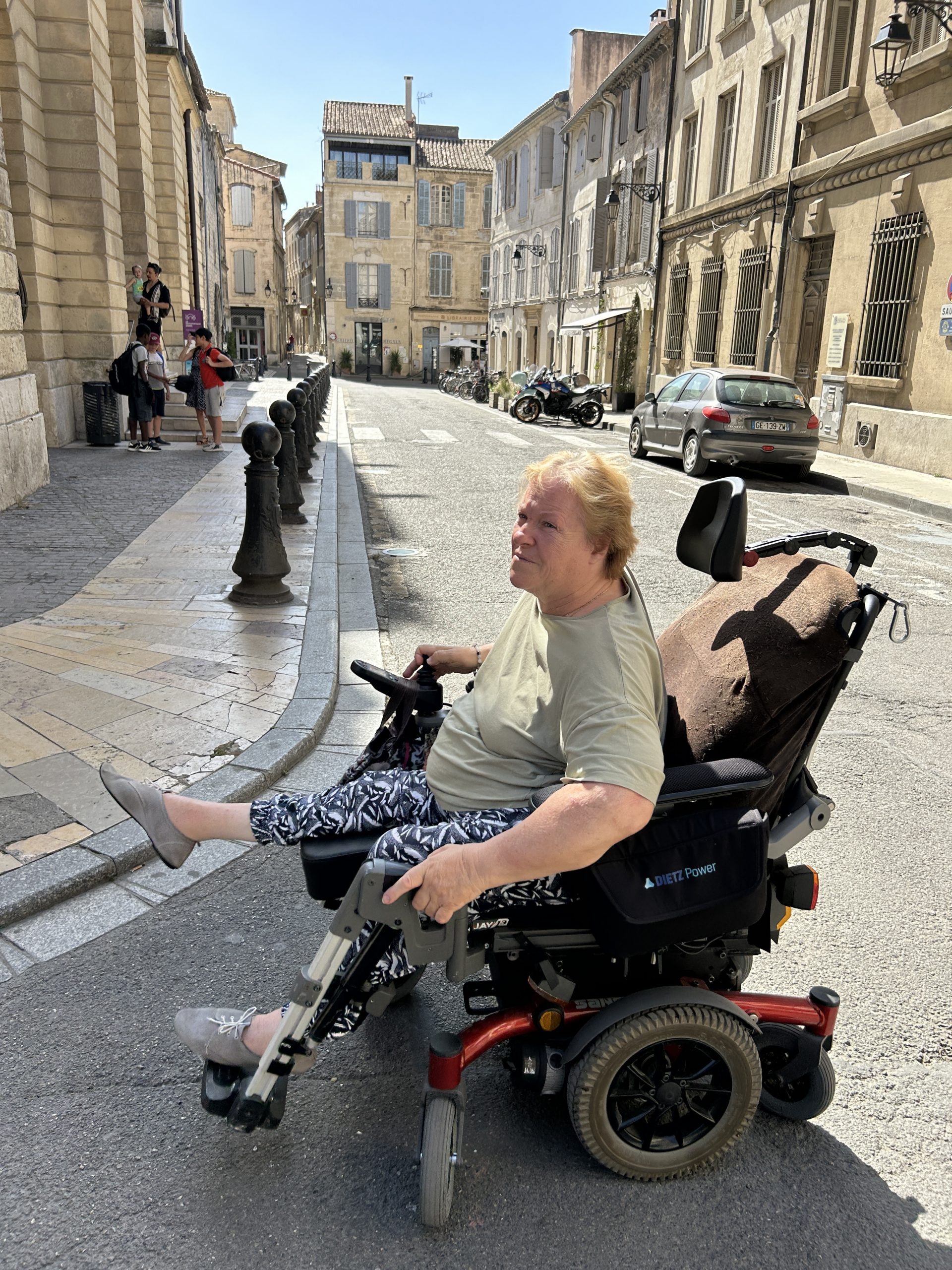 Woman in a wheelchair maneuvers through the a street in Arles, looking at a sidewalk without a curb cut.