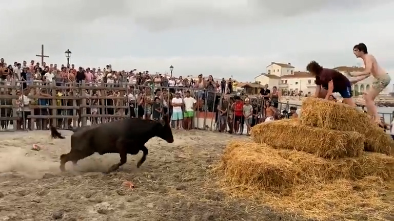 Two men stand on a haystack as a bull charges toward them.