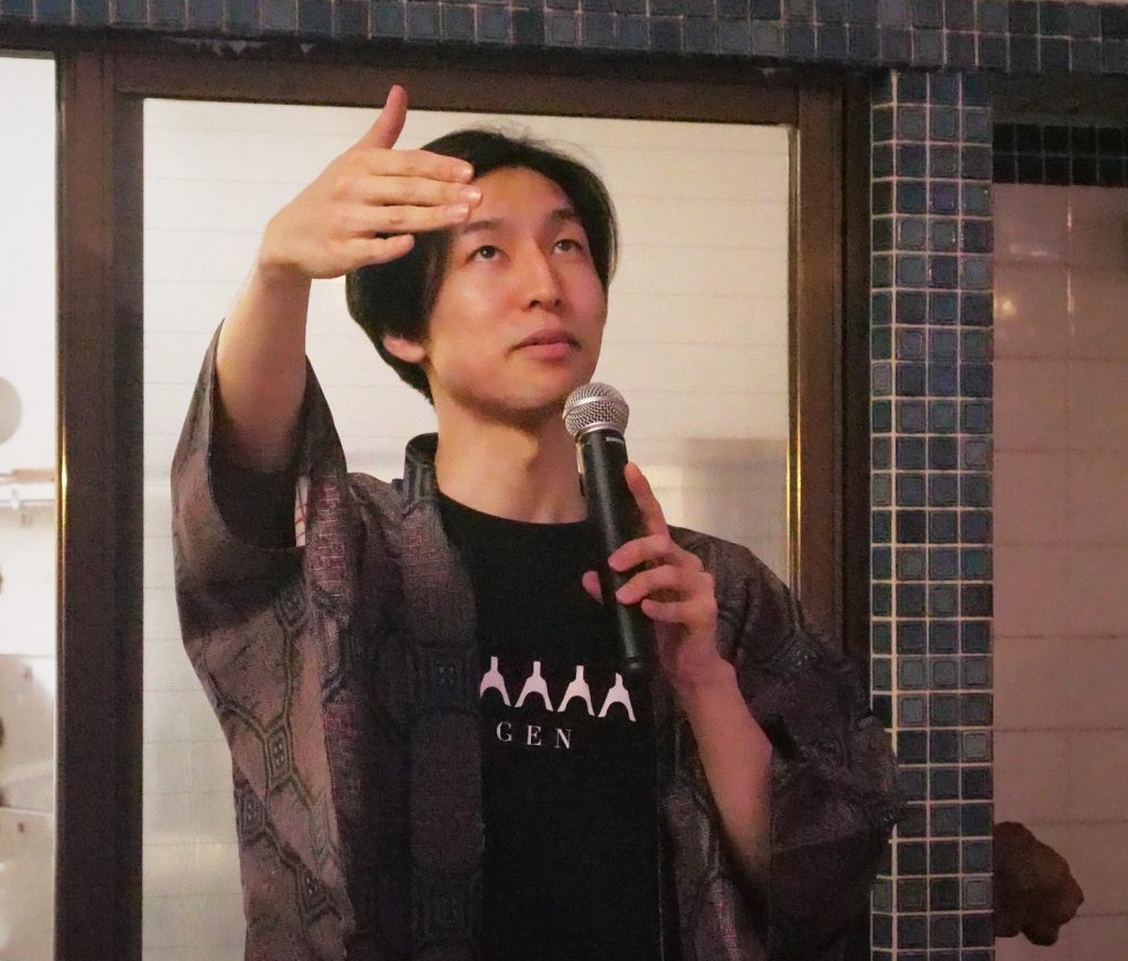 Concert organizer Shoehi Higashi raises one arm and holds a microphone with the other.