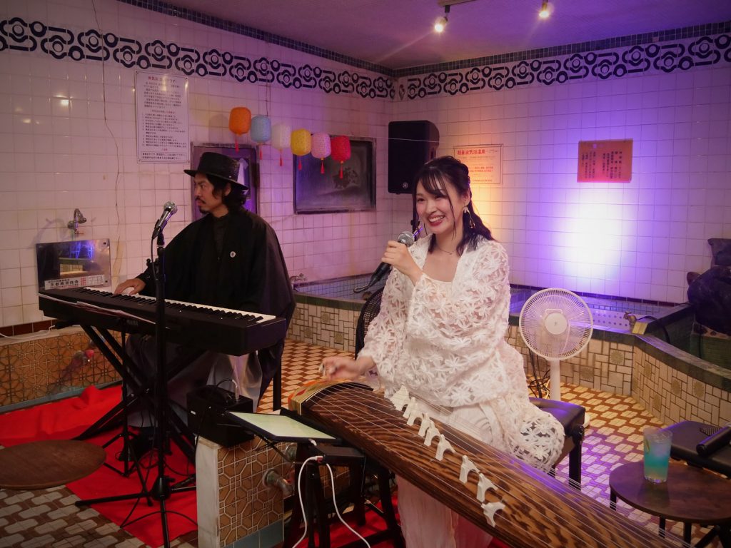 A woman in a white dress and a man perform music in an old Japanese sentō.