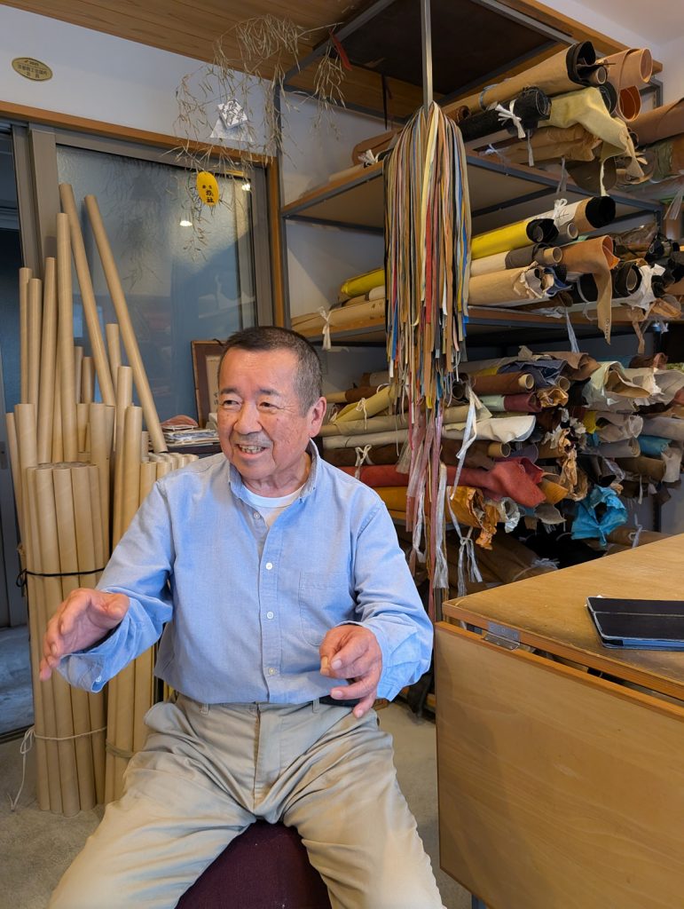 A man sits in a leather shop with rolls of leather behind him