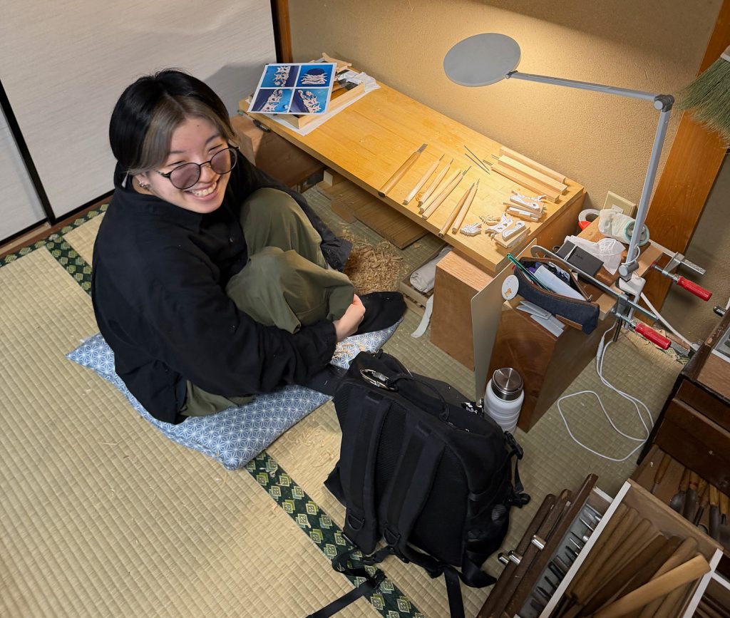 A young woman sits on a tatami mat-covered floor at a workbench.