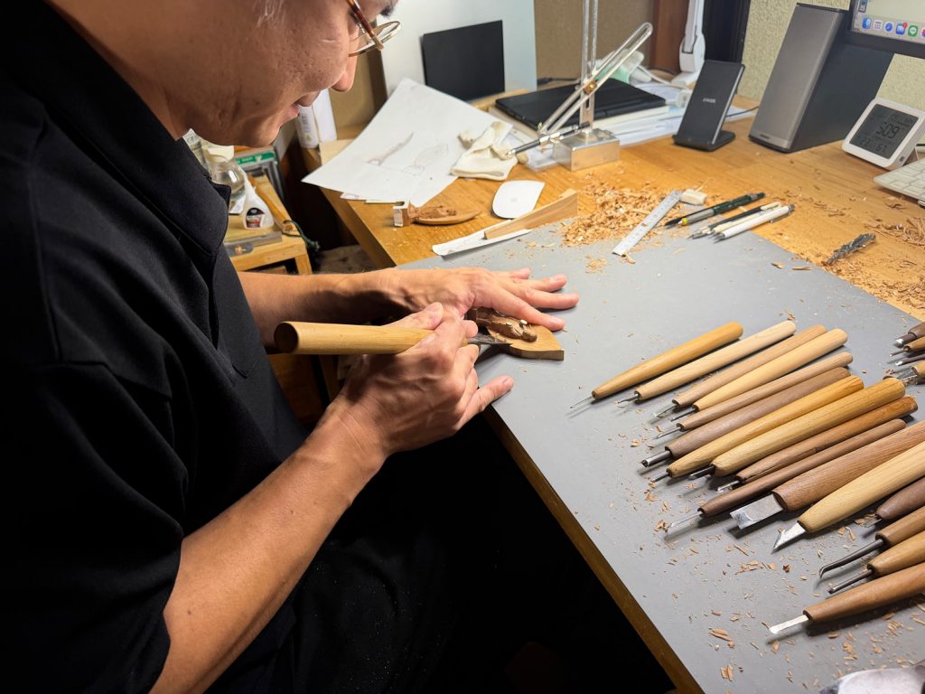 A woodcarver carves a tiny Buddha as he leans over a table full of carving tools. Photo by Emma Cooney.