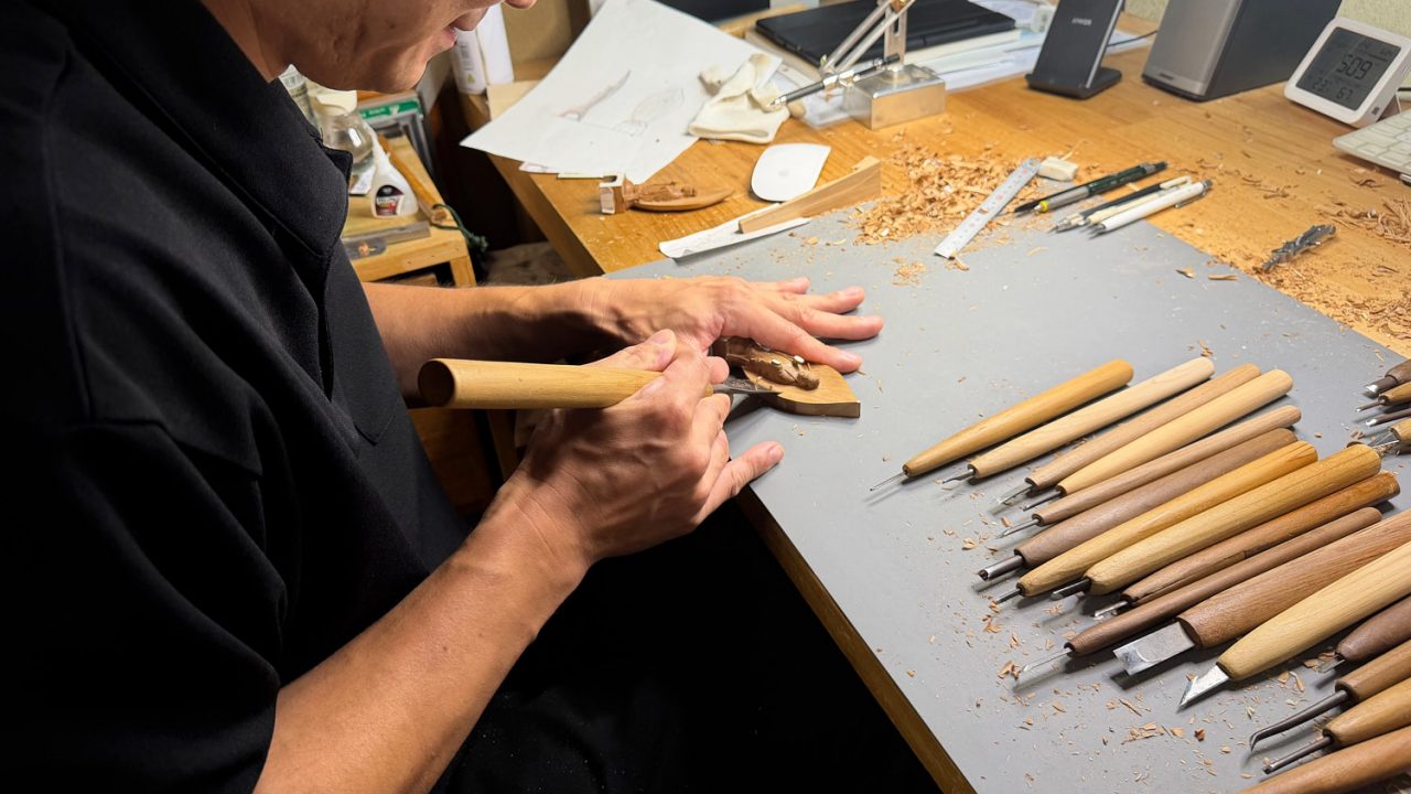 A woodcarver carves a tiny Buddha as he leans over a table full of carving tools. Photo by Emma Cooney.