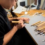 A woodcarver carves a tiny Buddha as he leans over a table full of carving tools. Photo by Emma Cooney.
