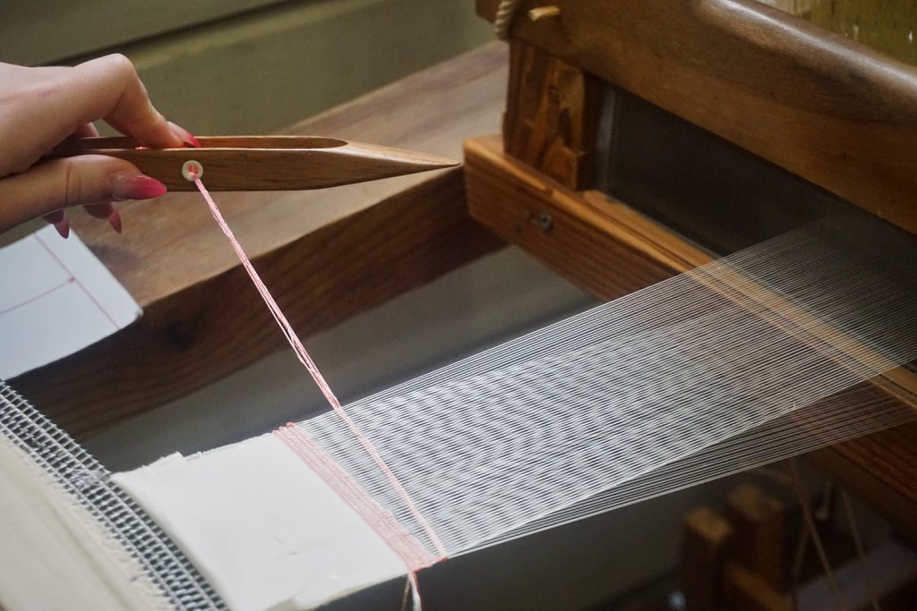 A wooden shuttle carries pale pink thread across the loom, adding another line to a piece of handwoven cloth. Photo by Katherine Istomin
